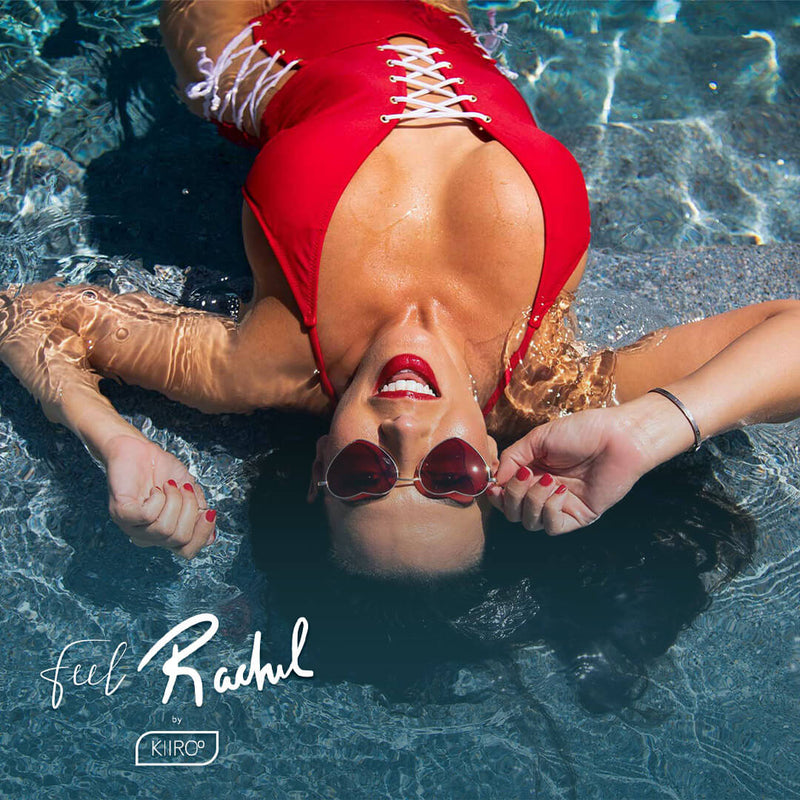 Rachel Starr is wearing a red, one-piece swimsuit and laying in a pool in the bright sunlight. The camera is above her, pointed down at her. She smiles at the camera, touching her heart-shaped sunglasses in a playful expression. | Kinkly Shop