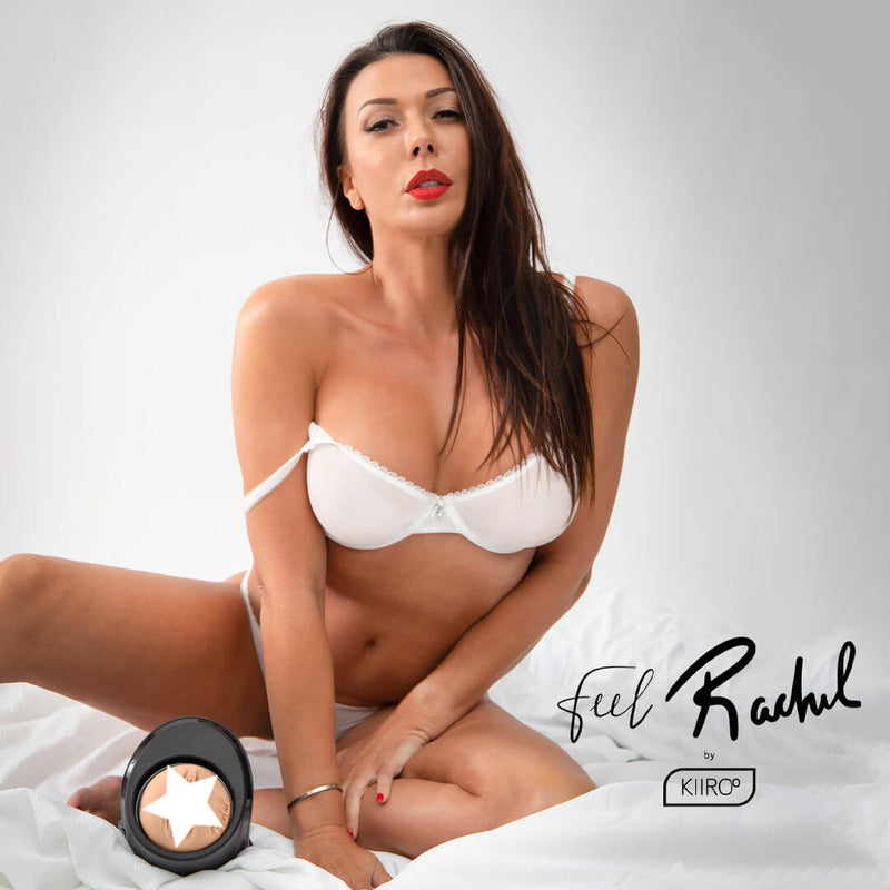 Rachel Starr in a white bra and panty set sitting in a white bedroom set. She stars into the camera seductively with one of her bra straps sliding down her arm. The KIIROO FeelStars FeelRachel Stroker, inserted in the KEON, is pointed towards the camera and resting between her legs. | Kinkly Shop