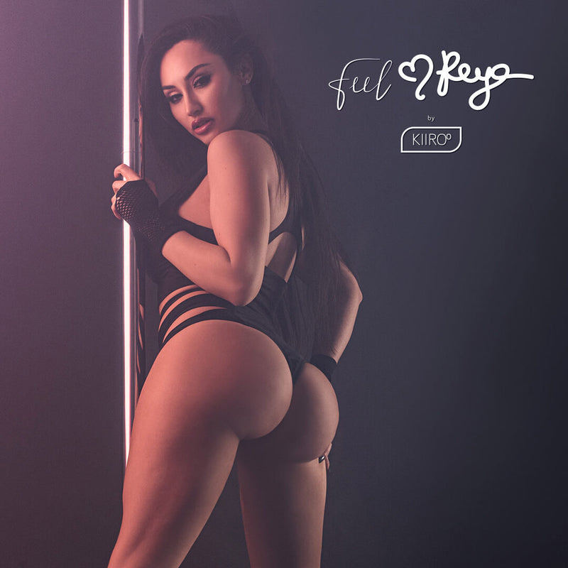 Reya Sunshine in black strappy workout clothing and fishnet gloves holding onto a stripper dance pole. Her butt is towards the camera, and she's look seductively into the camera. | Kinkly Shop