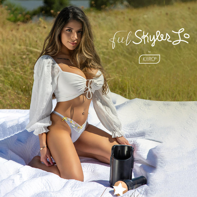 Skyler Lo kneeling on a white picnic blanket in a field. She's wearing a white croptop that displays a lot of cleavage as well as a thong. The KEON and the KIIROO FeelStars FeelSkyler Stroker are laying out in front of her. | Kinkly Shop