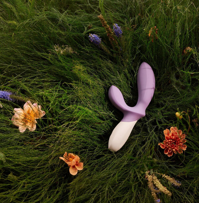 The LELO LOKI Wave in violet laying out in a field of long green grass. There are flowers all around the toy. | Kinkly Shop