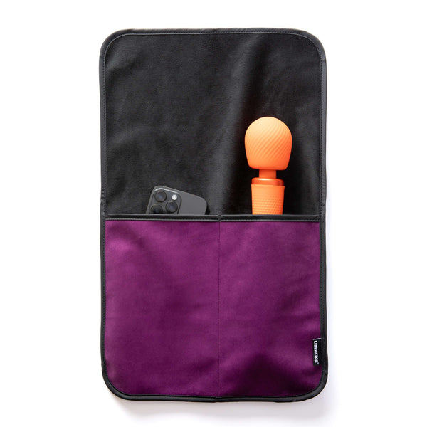 The Liberator Fascinator Sex Toy Caddy in Purple laying flat on a white surface. A wand massager and a cell phone are shown peeking out from the tops of the pockets. The full length of the wand massager fits within the deep pocket, leaving only the massager's head showing from the pocket. | Kinkly Shop