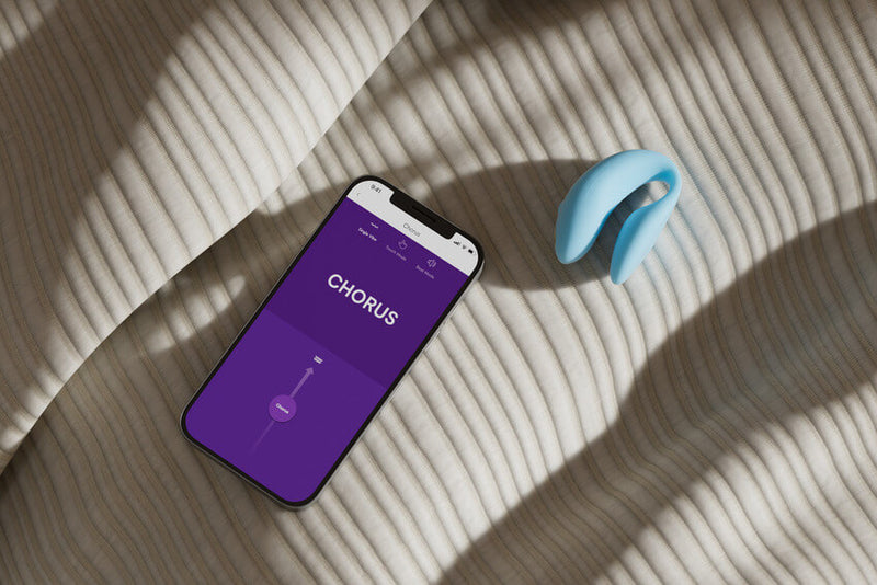 The We-Vibe Chorus lays out on a textured bedspread next to a cell phone. The cell phone is open to the We-Vibe Connect app, showcasing the slider control that would allow the phone to control the vibrator. | Kinkly Shop