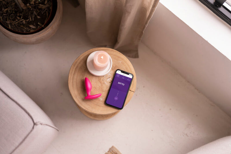 The We-Vibe Ditto+ in Cosmic Pink sitting on a wooden end table. A lit candle and a cell phone, connected to and controlling the We-Vibe Ditto+, are shown next to it. | Kinkly Shop