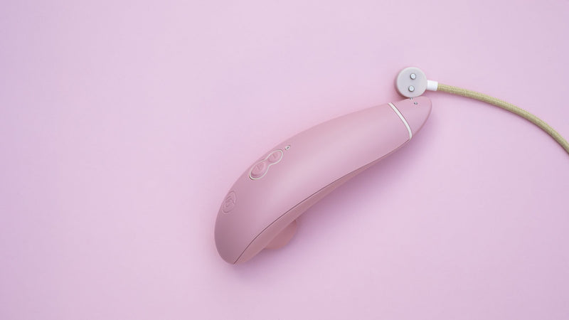 The Womanizer Premium Eco is laying against a pink background with the round, magnetic charging laying next to the vibrator's magnetic charging port. | Kinkly Shop