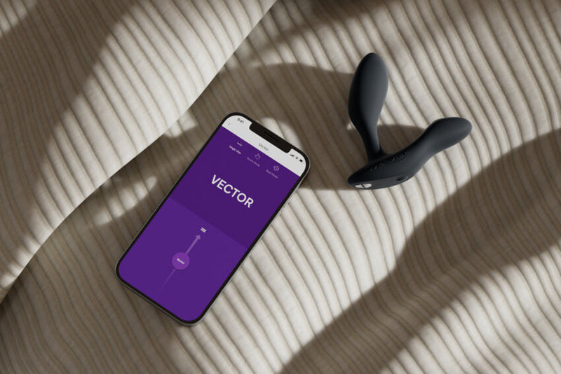 The We-Vibe Vector+ in black sitting on bedsheets. A cell phone is sitting next to it with the We-Vibe app open and currently controlling the sensations on the Vector+ | Kinkly Shop