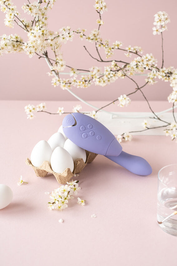 Womanizer Duo 2 in Lilac lays out against a pink background. There are white cherry blossom-like flowers in the background. The Duo 2 is resting up against a six-pack carton of eggs. It is noticeably much larger than an egg. | Kinkly Shop