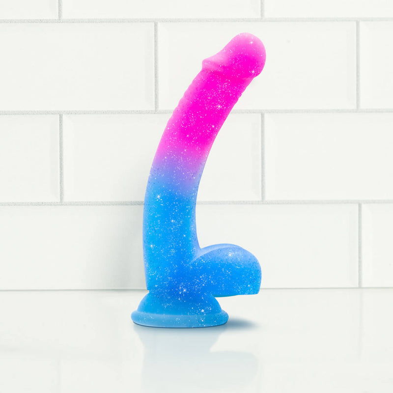 The Blush Avant Chasing Sunsets dildo up against a white tiled wall. The dildo has a noticable curve to it alongside a pair of testicles near the base. Underneath the testicle base is a large suction cup. | Kinkly Shop
