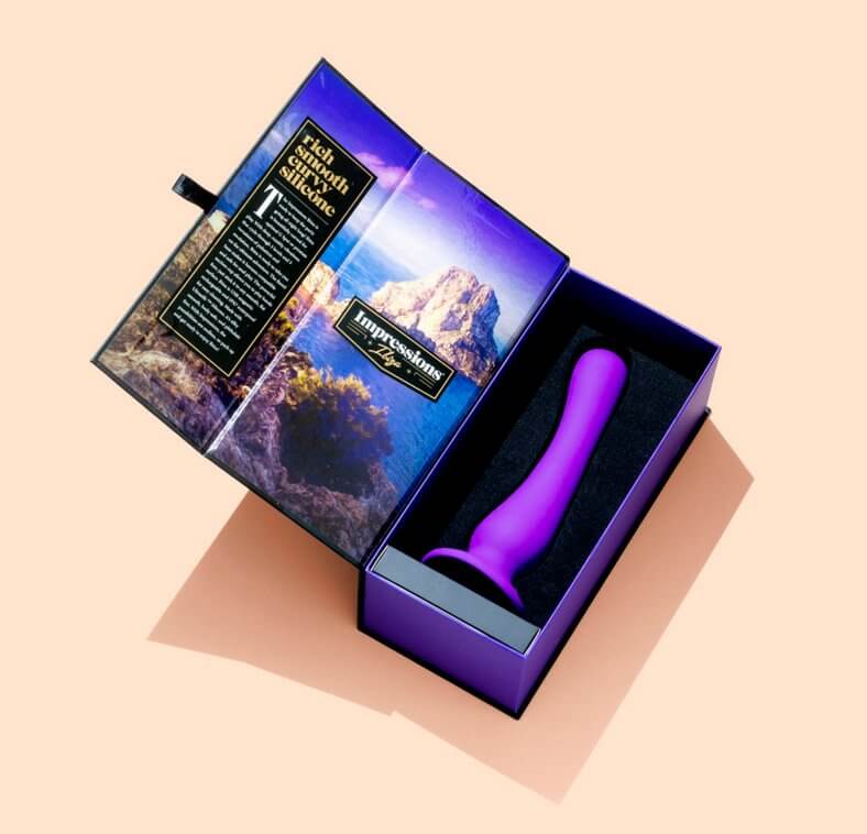 The Blush Impressions Ibiza dildo is resting inside of the foam packaging. The packaging is open and displaying the landscape photo the sex toy is named after. | Kinkly Shop