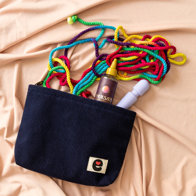 The Emojibator Pleasure Pouch laying on top of peach sheets on a bed. The pouch is unzipped, and items are spilling out from it. It has a hank of colorful rope, a bottle of lube, and a mini wand massager inside of it. | Kinkly Shop