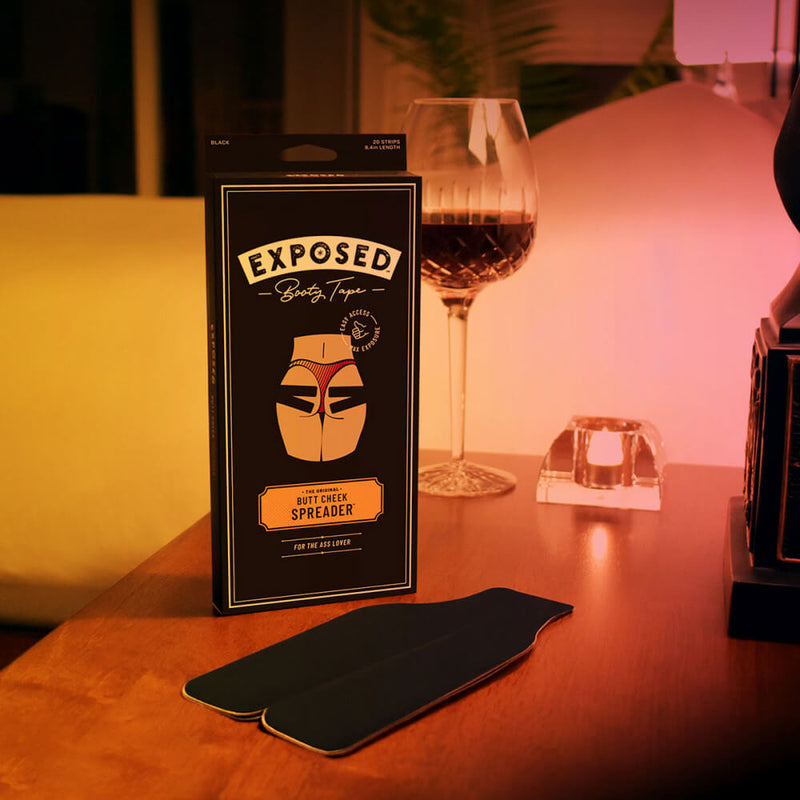 The Exposed Booty Tape is laying on a table in front of the packaging for the Exposed Booty Tape. A wine glass is in the background, and the entire photo is dimly lit - implying a sensual night in. | Kinkly Shop