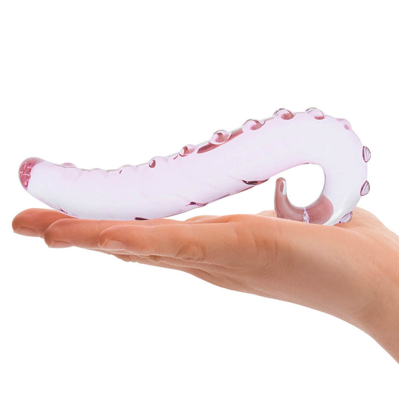 A palm laid out flat in front of a white background. The Glas Lick-It Glass Dildo rests on top of the open palm. This showcases the upward curve of the tip of the dildo as well as the easy-grip looped base. The dildo is also clearly translucent, and the white background can be seen through the body of the toy. | Kinkly Shop