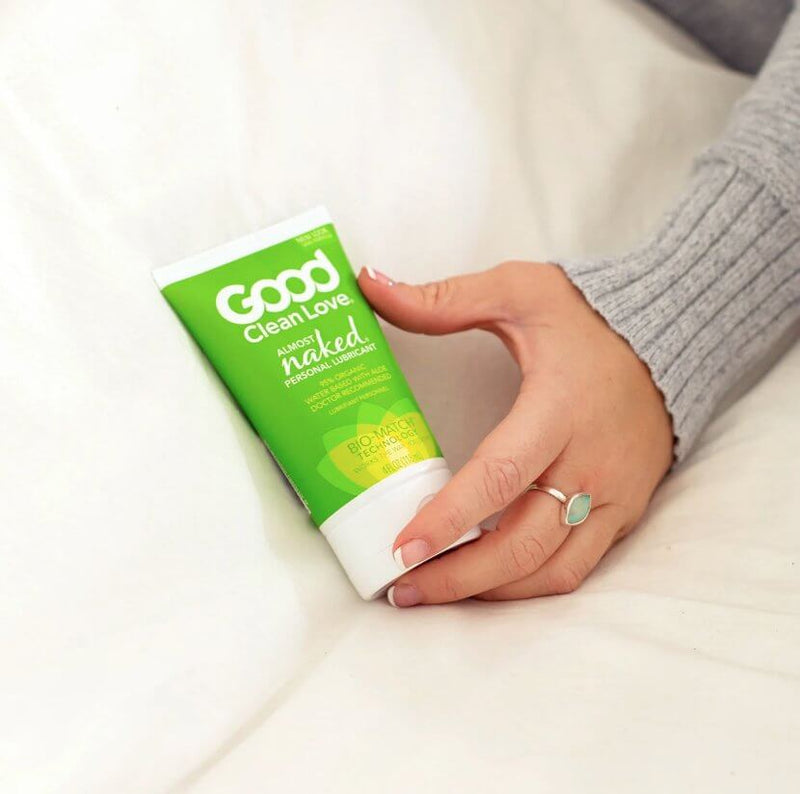 A hand holds the Good Clean Love Almost Naked Organic Lubricant - 4OZ on a bedspread. The bottle is taller than the space between the person's pointer finger and their thumb, and it looks like it would be about the length of their hand from fingertip to base of the palm. | Kinkly Shop
