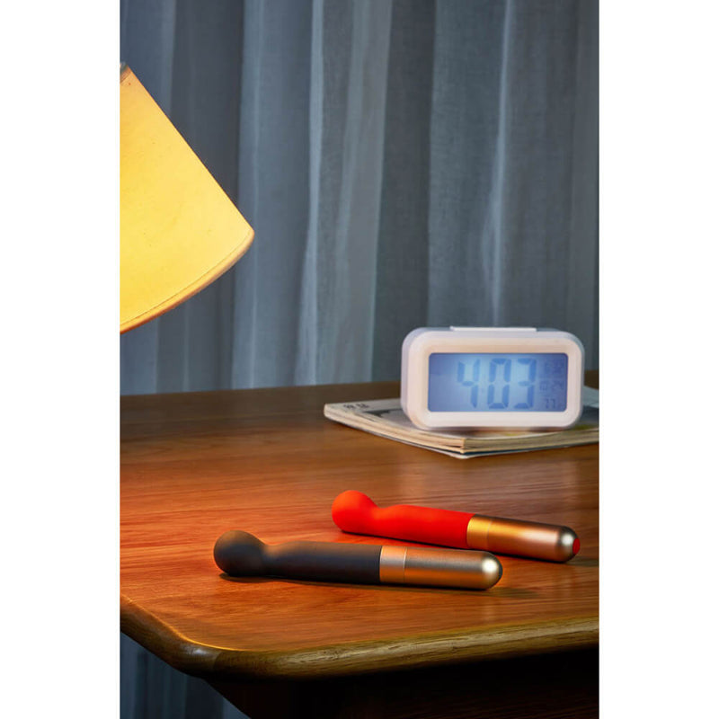 The Love Not War Kama in Orange and Grey sit out on top of a wooden bedside table. A bedside lamp is shining a warm, yellow "spotlight" onto the two vibes. Sheer white curtains are shown in the background along with a digital alarm clock that reads "4:03". | Kinkly Shop