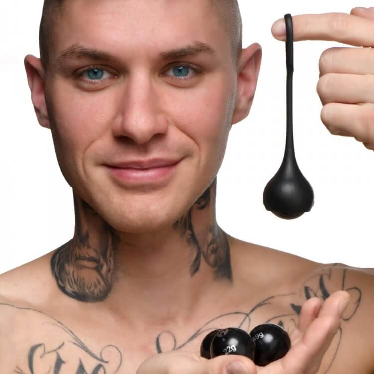 A person with tattoos is looking at the camera and smiling. The Master Series Cock Dangler penis weights is hanging from their pointer finger. | Kinkly Shop