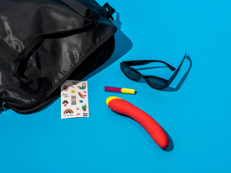 The ROMP Hype G-Spot Vibrator sitting out next to an open bag. The ROMP Hype G-Spot Vibrator is surrounded by a pair of sunglasses, a lip gloss, and a set of ROMP stickers. The vibrator is much thicker than the lip gloss tube and is longer in length than the sunglasses. | Kinkly Shop
