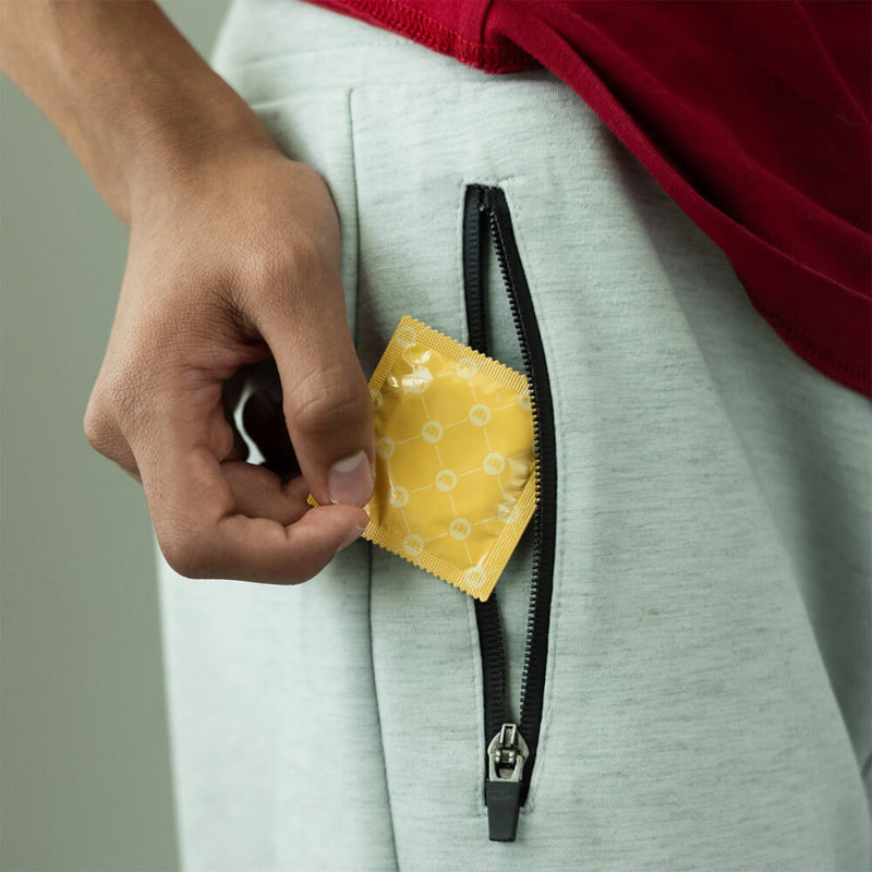 A person in grey sweatpants is tucking a Royal Tailored Fit Ultra Thin Vegan Latex Condom into their zippered pocket. The condom is bright yellow with no lettering on it. | Kinkly Shop