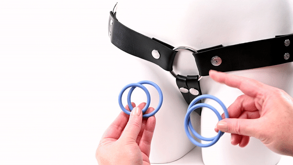 GIF shows a minimalist strap-on harness. A hand holds the four Sportsheets O-Ring 4-Pack rings - and in the next shot, one of the rings is shown strapped into the harness. The text on the GIF reads "Sizes: 1.5", 1.75", 2", and 2.25". | Kinkly Shop
