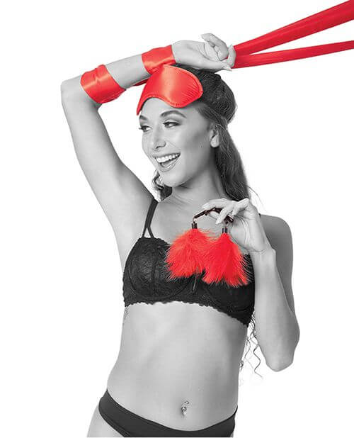 A person is smiling and laughing as they wear the red blindfold, have the red satin sash wrapped around their wrists, and holding the two feathered nipple clamps in their hands. | Kinkly Shop