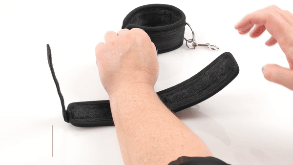 GIF that shows a person handling the Sportsheets Soft Wrist Cuffs. The person fastens themselves into a cuff and fastens it with the velcro onto their wrist. They then tug onto the cuffs to show the durability. The text says "Starter Restraints" and "For couples interested in fantasy play." | Kinkly Shop