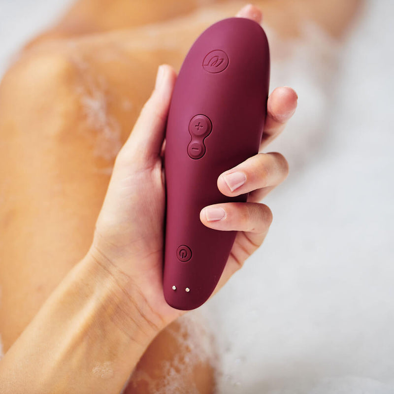 A hand holds the Womanizer Classic 2 in Bordeaux. In the background, the image shows the bare person's smooth legs and the white fluffiness of bath bubbles. The Bordeaux's strong color is stunning in contrast. | Kinkly Shop