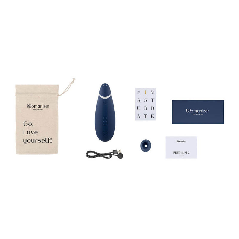 The Womanizer Premium 2 sitting out on a white background with everything that comes with the air suction vibrator. This includes the vibrator itself, a branded drawstring pouch, the charging cable, safety information, the instruction manual, warranty information, and an extra attachment head in a different size. | Kinkly Shop