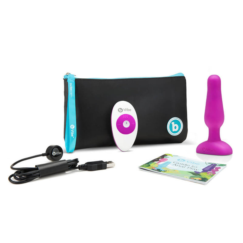 The b-Vibe Remote Novice Plug in Fuchsia shown with everything that the plug comes with. This includes the plug itself, the remote, the Guide to Anal Play, the charging cable, and the branded, zippered pouch. | Kinkly Shop