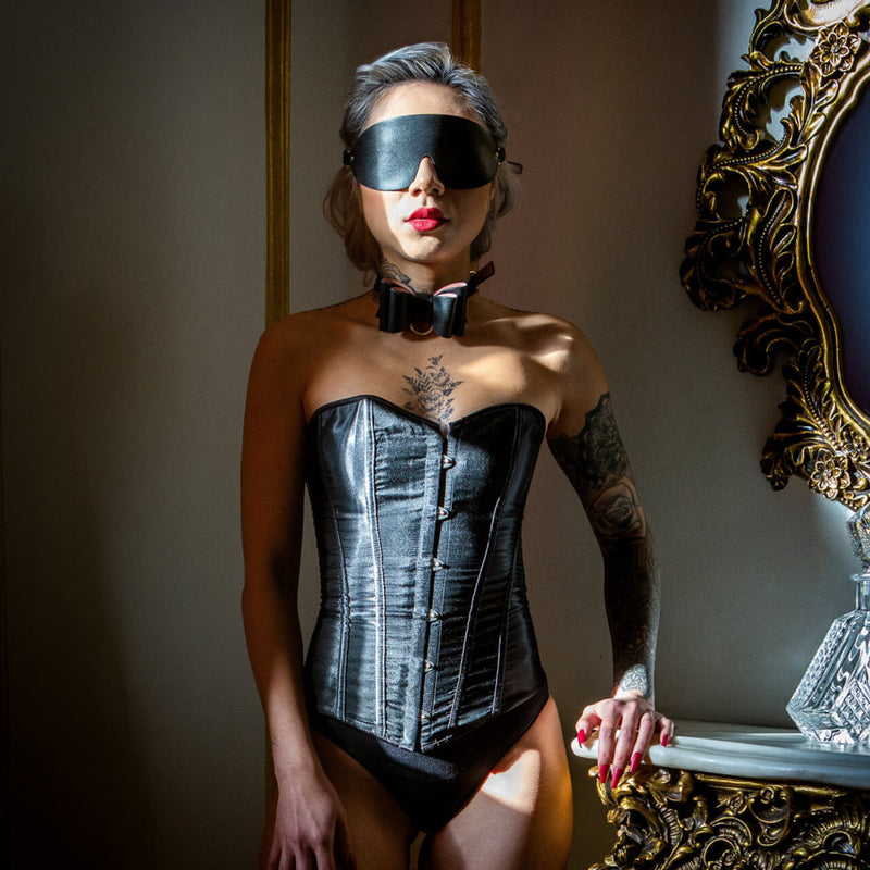 A person stands next to a dresser and a mirrow in a corset. They're wearing the blindfold and collar from the Blush Temptasia Safe Word Bondage Kit with a serene expression on their face. | Kinkly Shop