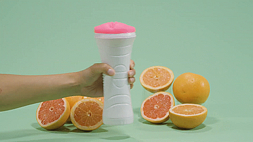 GIF of the Clone-A-Pussy Plus+ Sleeve. A person is holding it in their hand in front of an arrangement of oranges. They hold the Clone-A-Pussy Plus+ Sleeve upright then tilt it towards the camera to showcase the cloned vulva at the entrance of the sleeve. They use a finger to pull at one of the inner labia of the cloned vulva. | Kinkly Shop