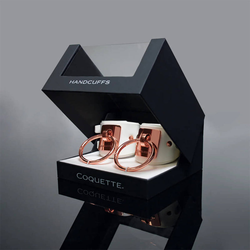 The Coquette White and Rose Gold Adjustable Cuffs inside of their packaging. They come in a black and see-through box that opens upwards like a luxury item. | Kinkly Shop