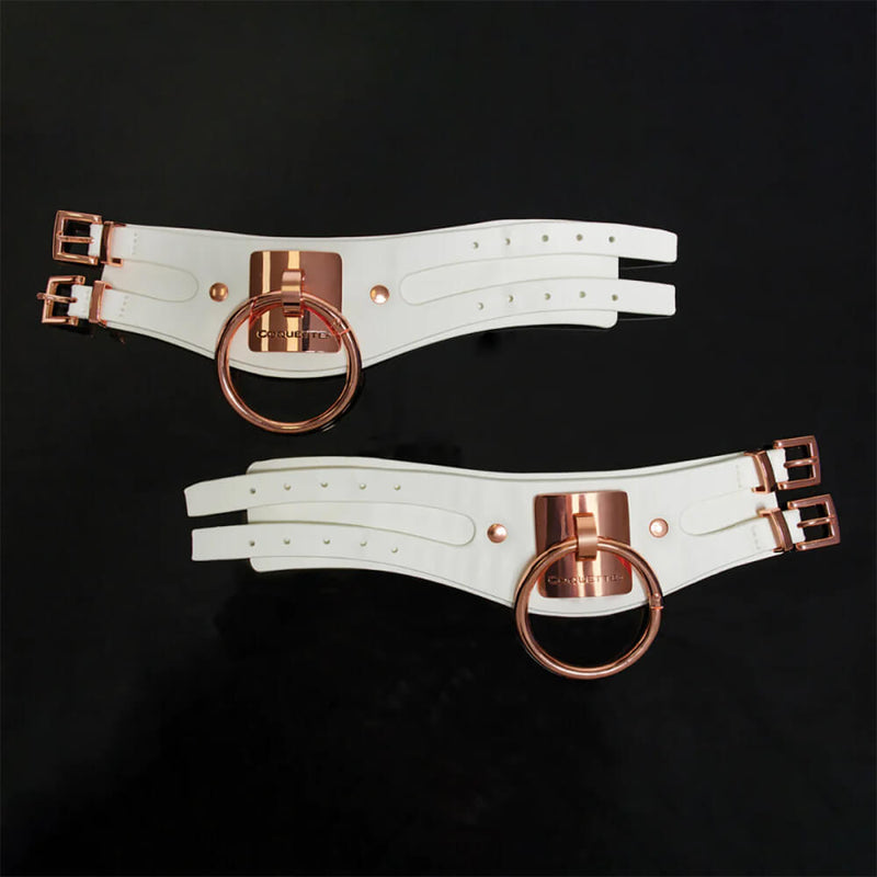 Both of the Coquette White and Rose Gold Adjustable Cuffs are laid flat on a black background. The cuffs are noticeably wider than most standard bondage cuffs with an extra-large, visually-catching O-ring on the front. They use two straps to fasten the cuffs onto the wrists. They are widest at the front of the cuff (with the O-ring plate) and get slightly narrower towards the side with the two fastening straps. | Kinkly Shop