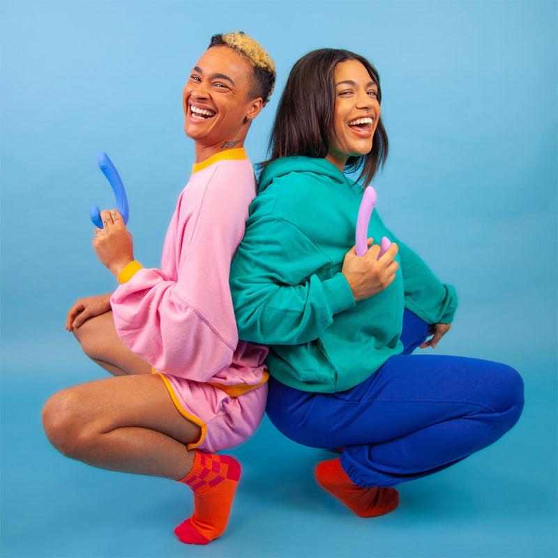 Two people are smiling and squatting in front of a blue background. Each person holds a Cute Little Fuckers Jix in a different color. Their grips look comfortable, and the Jix protrudes away from their hands with a long, slender length. | Kinkly Shop
