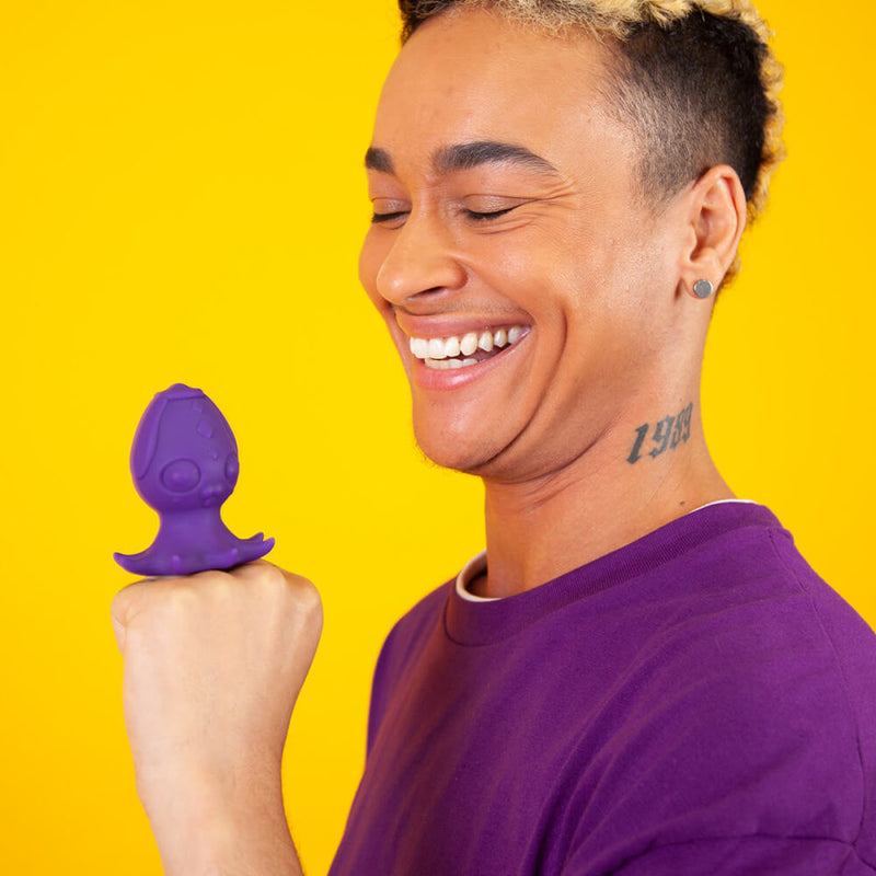 A person is smiling in front of a bright yellow background. They are making a fist with the "flat" portion of their hand pointing upwards. A purple Cute Little Fuckers Princette Puppypus rests on top of their fist, showcasing the toy's flat, sit-upright base. | Kinkly Shop