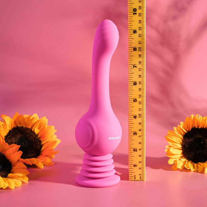 The Evolved Gyro Vibe with a pink background, surrounded by sunflowers. A yellow construction ruler is placed up against the Evolved Gyro Vibe to showcase how tall it is. The Evolved Gyro Vibe comes up to a little over 9.5" in height. All measurements of the toy are listed within the text description for accessibility. | Kinkly Shop