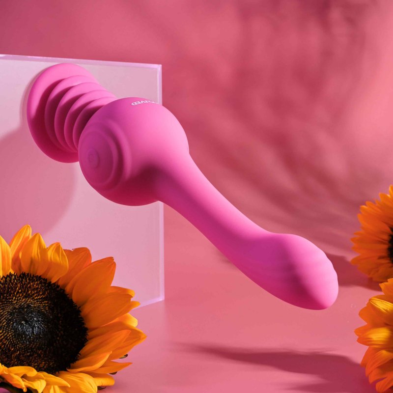 The Evolved Gyro Vibe hanging horizontally because the base is suction cupped up against a nearby, opaque box. The entire scenery is pink, matching the pink Evolved Gyro Vibe, with sunflowers scattered around in the foreground for visual interest. The toy is hanging, fully, with no support due to the powerful suction cup base. | Kinkly Shop