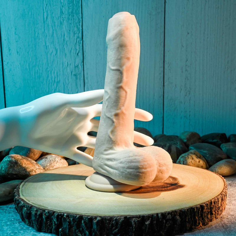 The Evolved Peek a Boo Vibrating Dildo sits out on a flat piece of wood. A mannequin hand goes to reach around it. It looks like the hand could comfortably wrap its fingers around the shaft of the dildo. | Kinkly Shop