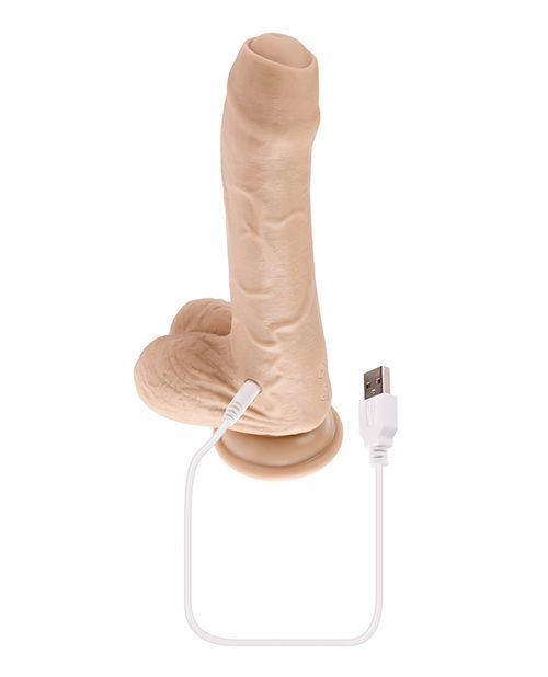 The Evolved Peek a Boo Vibrating Dildo sitting upright against a white background with the charging cable plugged into the base. The charging port is near the base of the shaft, by the testicles. | Kinkly Shop