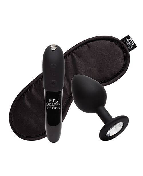 Everything included with the Fifty Shades of Grey Come to Bed Kit sits against a white background. There's the We-Vibe Tango X bullet vibrator in a black Fifty Shades of Grey design, a black butt plug with a circular, gem-shaped base, and a black, satin, branded Fifty Shades of Grey blindfold. | Kinkly Shop