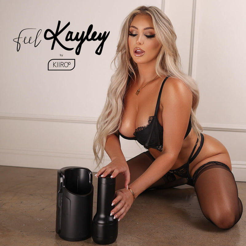 Kayley Gunner in black lingerie kneeling on the floor in front of a regal white wall. Her make-up is done immaculately. She's leaning forward to touch the KIIROO FeelStars FeelKayley Stroker next to the KEON. | Kinkly Shop