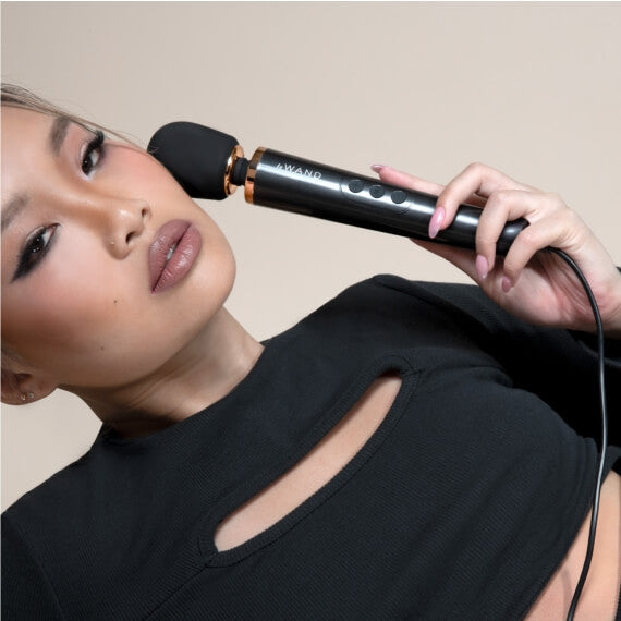 A person is holding the Le Wand Petite Plug-In Massager in black with the head up against their face. It looks like the head of the wand massager takes up about as much space as the person's lips. | Kinkly Shop