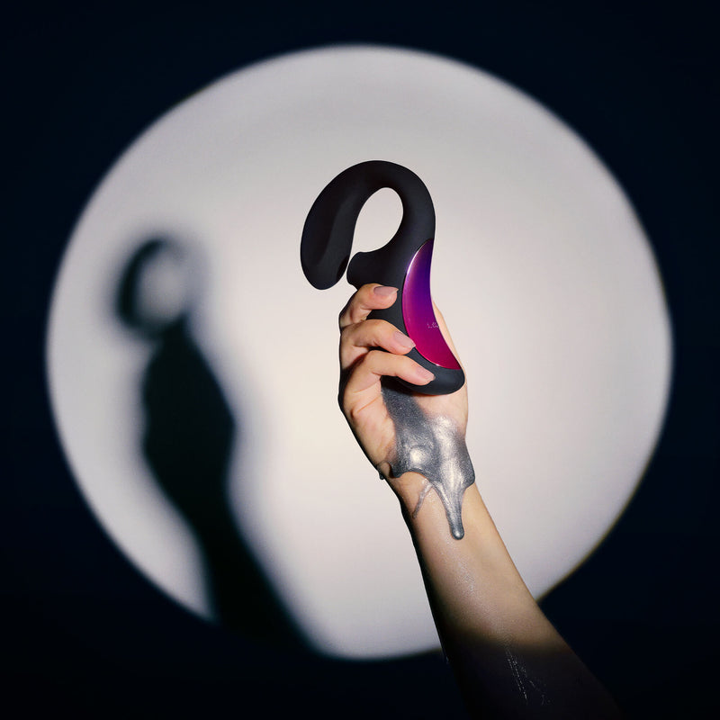 A spotlight shines on a hand holding the LELO Enigma. Silver paint drips down their wrist. The toy's clitoral end fits comfortably in the person's hand while the insertable shaft extends outwards past the toy. | Kinkly Shop