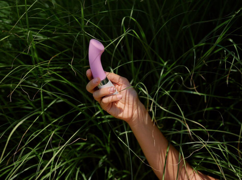 A hand holds the LELO GIGI 2 amid some tall grass. The vibrator is somewhat shadowed, but it stands out really well against the dark background of the green grass. | Kinkly Shop