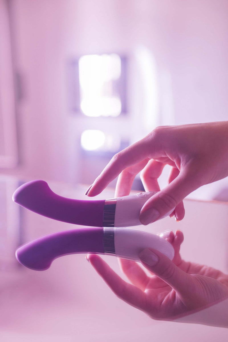 A hand goes to pick up the LELO GIGI 2 that's laying on a mirrored table. This side angle shows the deep g-spot curve that the Gigi 2 has. | Kinkly Shop