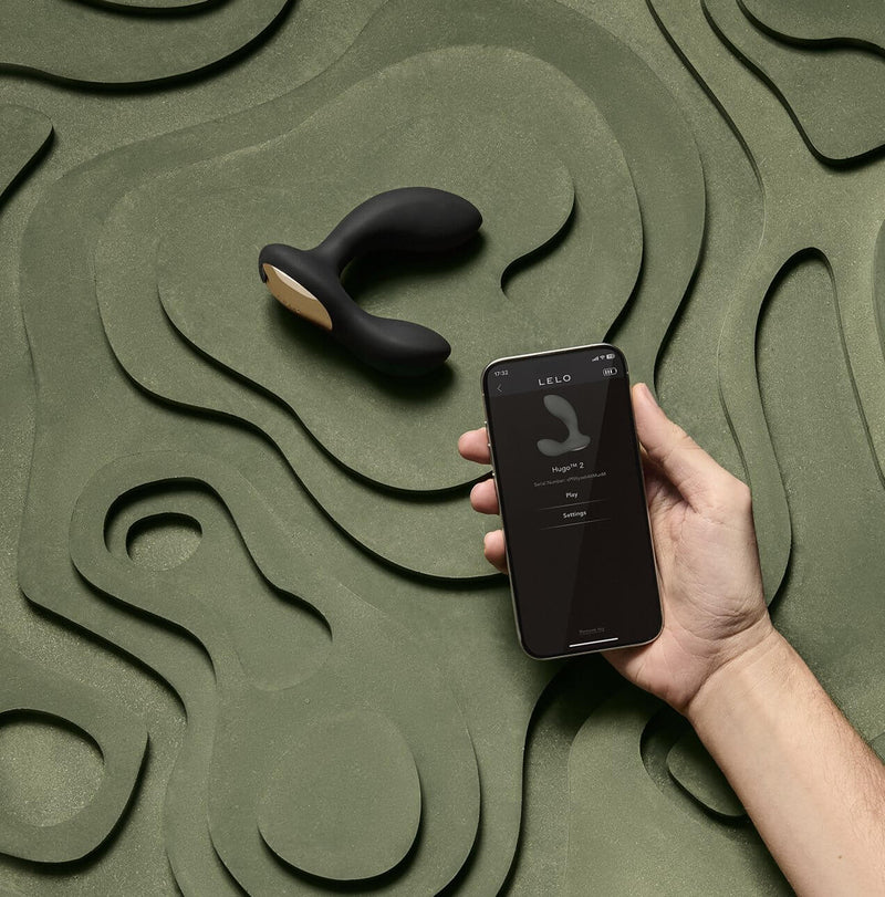 The LELO HUGO 2 without Remote lays out on a green textured surface. A hand is close to the camera, holding a cell phone with their cell phone open to the cell phone app, connecting the toy to the Hugo 2. | Kinkly Shop