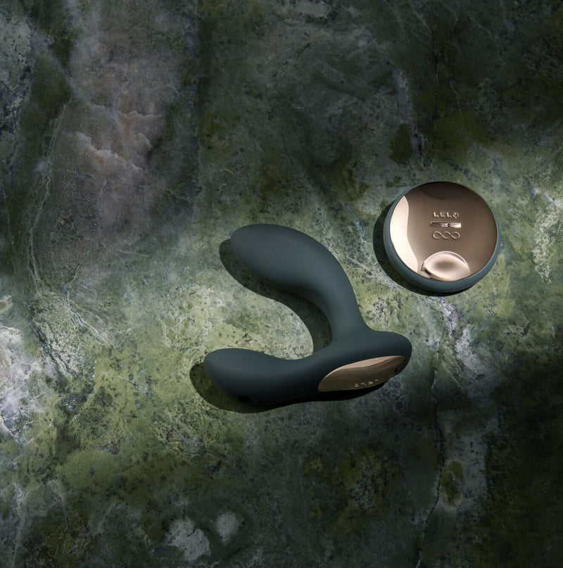 The LELO HUGO 2 in Black with Remote laying on top of a green marbled surface. A streak of light illuminates the toy and the remote, setting it apart from the shadowed areas around it. | Kinkly Shop
