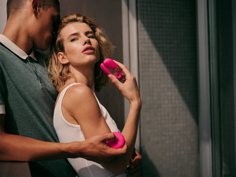A couple embraces near a window. One person is holding the remote in their hands, pressing a button. The other person is holding the vibrator in their hands, showcasing the slender, U-shaped design of the toy. | Kinkly Shop