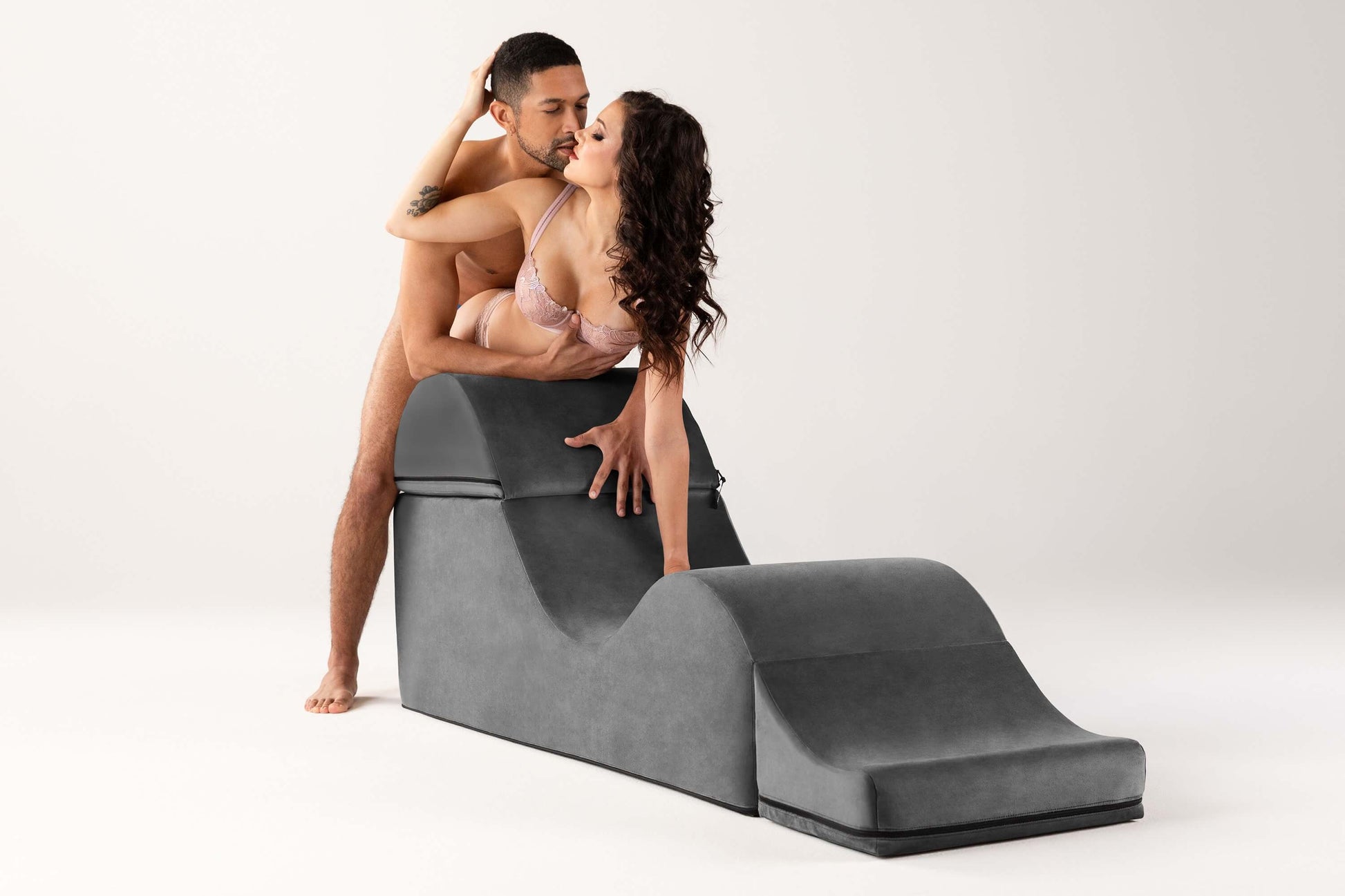 A couple embraces while using the Liberator Aria Flip Chaise. The receiving partner is standing at the tallest point, bent over at the waist overtop of the Aria Flip Chaise. The penetrating partner is standing behind them, pushing into them for penetration. | Kinkly Shop