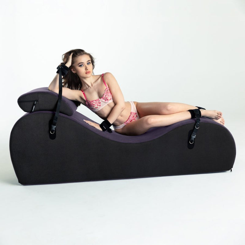 A person lounges on top of the Liberator Black Label Lyza Lounger Valkyrie. They are laying on their side. Despite having an elbow and hip supporting the majority of their weight, neither area is depressed underneath the person's limb. The person's wrists and ankles are restrained to the Liberator Black Label Lyza Lounger Valkyrie underneath them, and they're lounging on top of the included headrest shape. | Kinkly Shop