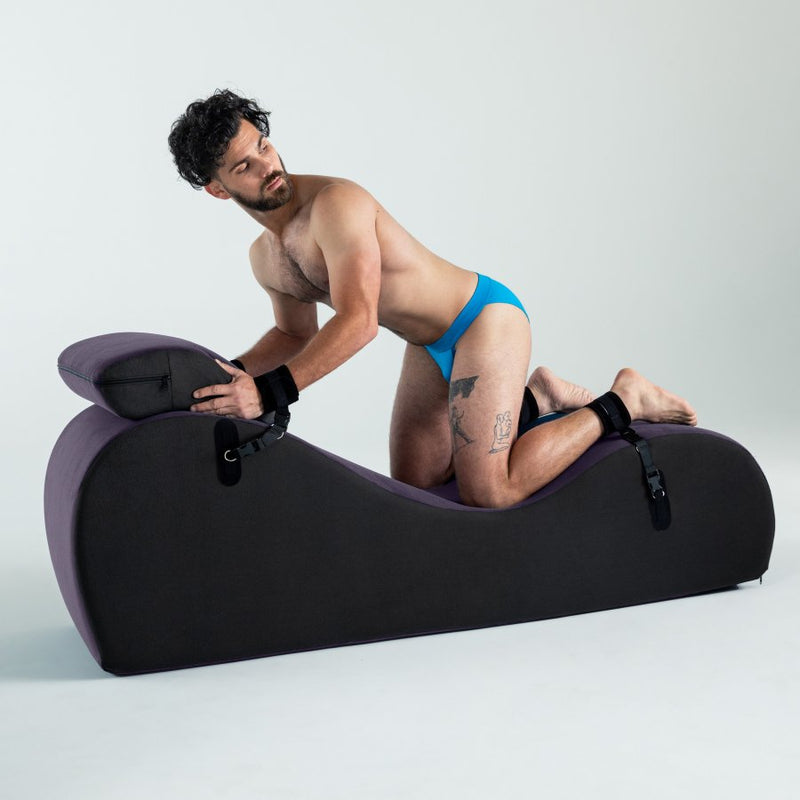 A person is kneeling on top of the Liberator Black Label Lyza Lounger Valkyrie, butt in the air, looking over their shoulder. The sex furniture is slightly compressed underneath the person's knees where all of their body weight is being held. Their wrists and ankles are restrained to the sex furniture underneath them. | Kinkly Shop