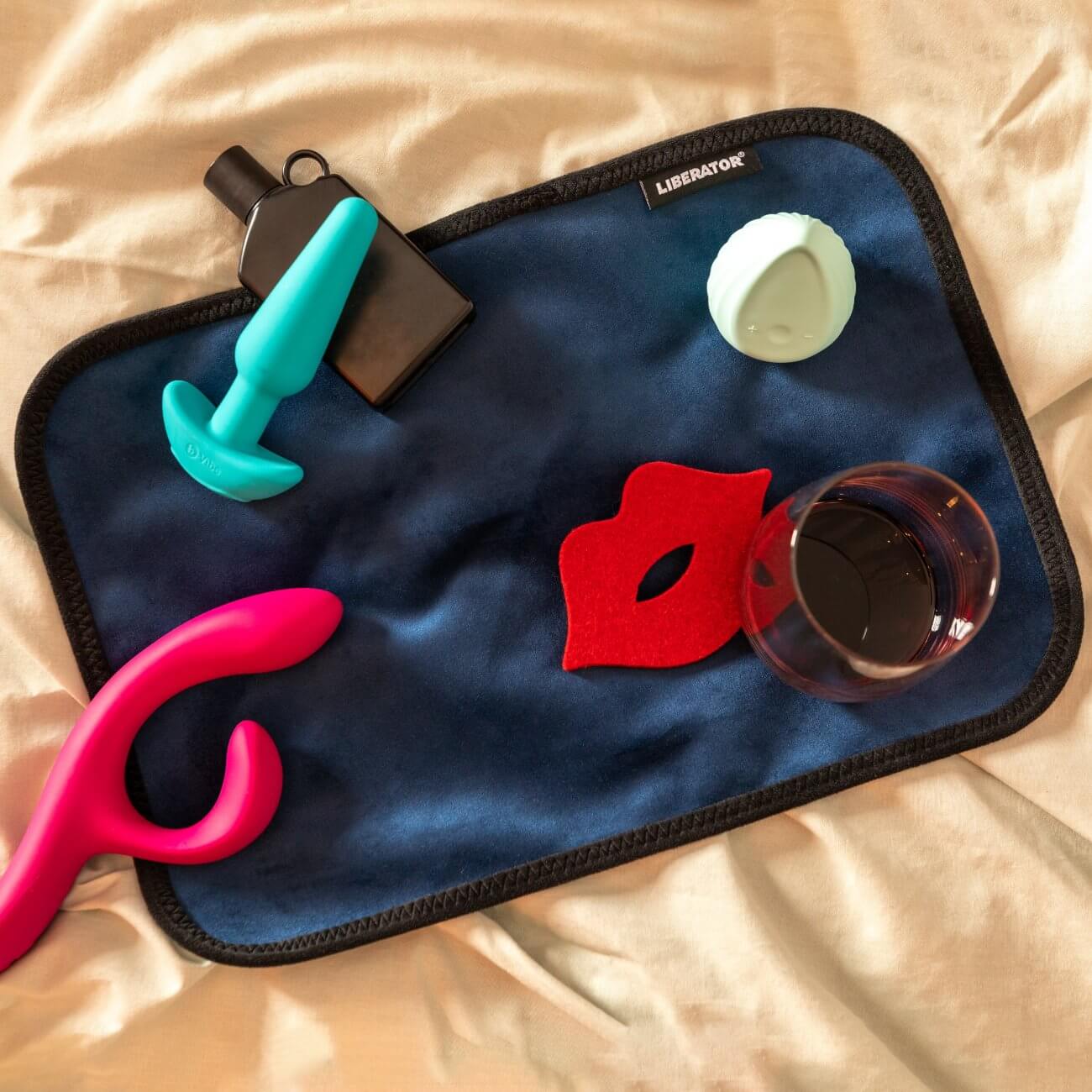 Three sex toys, a bottle of lube, a coaster, and a glass of wine all sit out on top of the Liberator Fascinator Toy Pad. There's still a ton of extra room for additional items. | Kinkly Shop